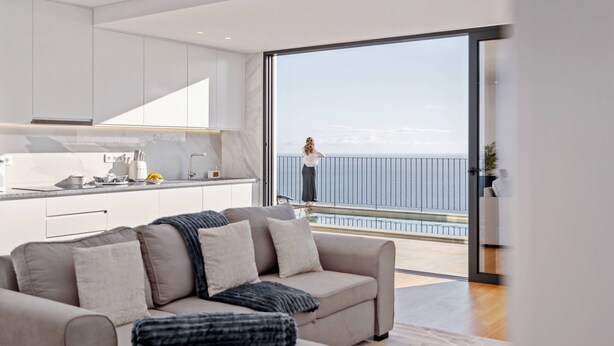 A modern living room with a sofa, a kitchen area, and a large sliding glass door leading to a balcony with a view of the sea. - Atlantic Bloom by Homie (Funchal)