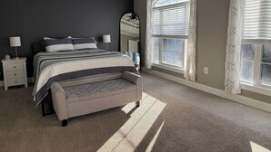 Een slaapkamer met een groot bed, een nachtkastje, een bankje, een spiegel en een raam met jaloezieën.
