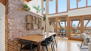 A dining area with a brick wall, wooden table, black chairs, and large windows. - Blue Mountain Retreat | Sunset Views | Fire Pit (Alexander)