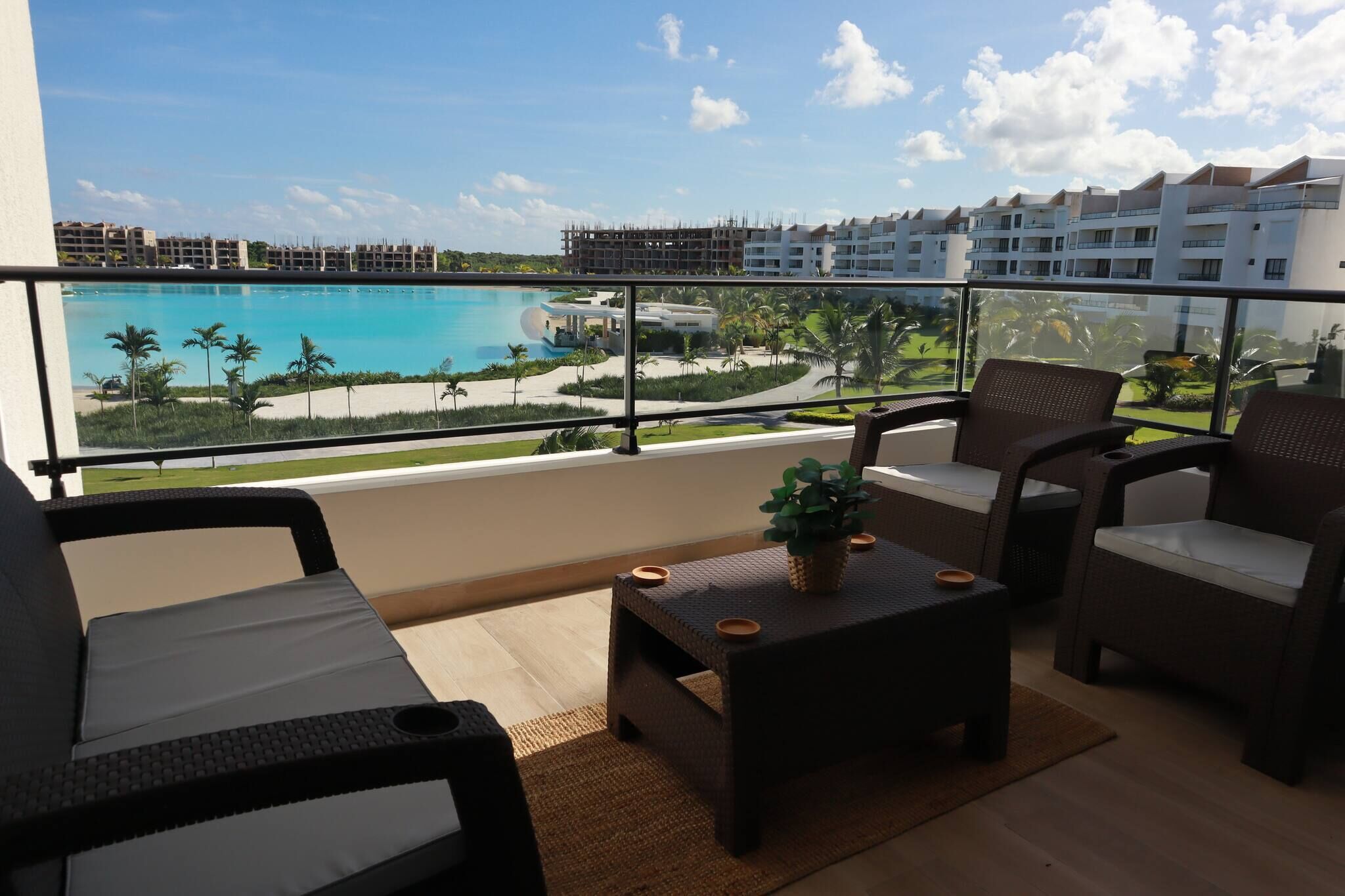 Un balcón con mobiliario de mimbre, una pequeña mesa con una planta y vistas a una piscina y edificios del complejo.