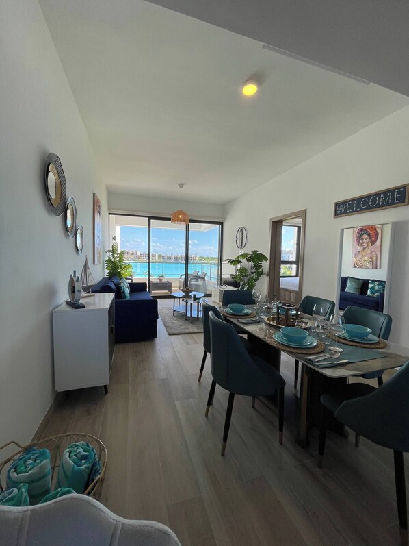 A modern dining area with a view of the sea, a living room with a sofa and a small table, and a 'WELCOME' sign on the wall. - Elegant Apartment in Thebeach (Punta Cana)