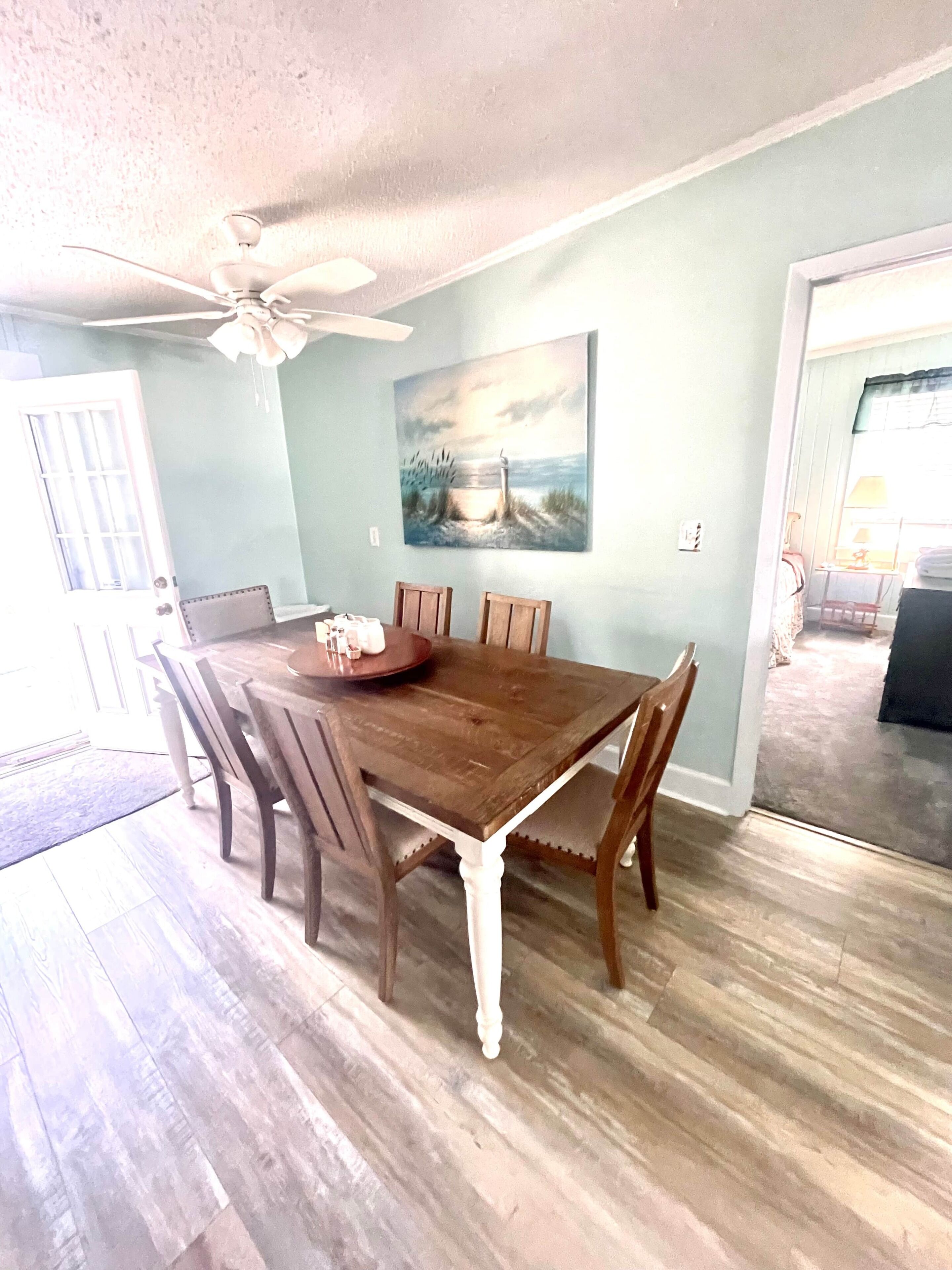 A dining room with a wooden table and chairs, a ceiling fan, and a painting on the wall.