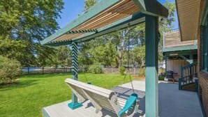 Um deck de madeira com baloiço, pérgola e vista para um quintal com relvado e árvores.
