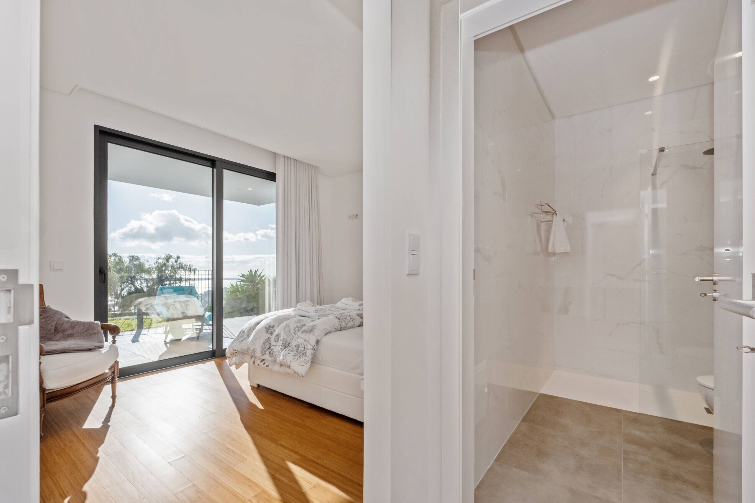 A modern bedroom with a large sliding glass door leading to a balcony, a separate shower area, and a wooden floor.