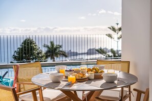A round table with breakfast items, including bowls, glasses, and fruits, on a balcony overlooking a pool and the ocean. - Atlantic Bloom by Homie (Funchal)