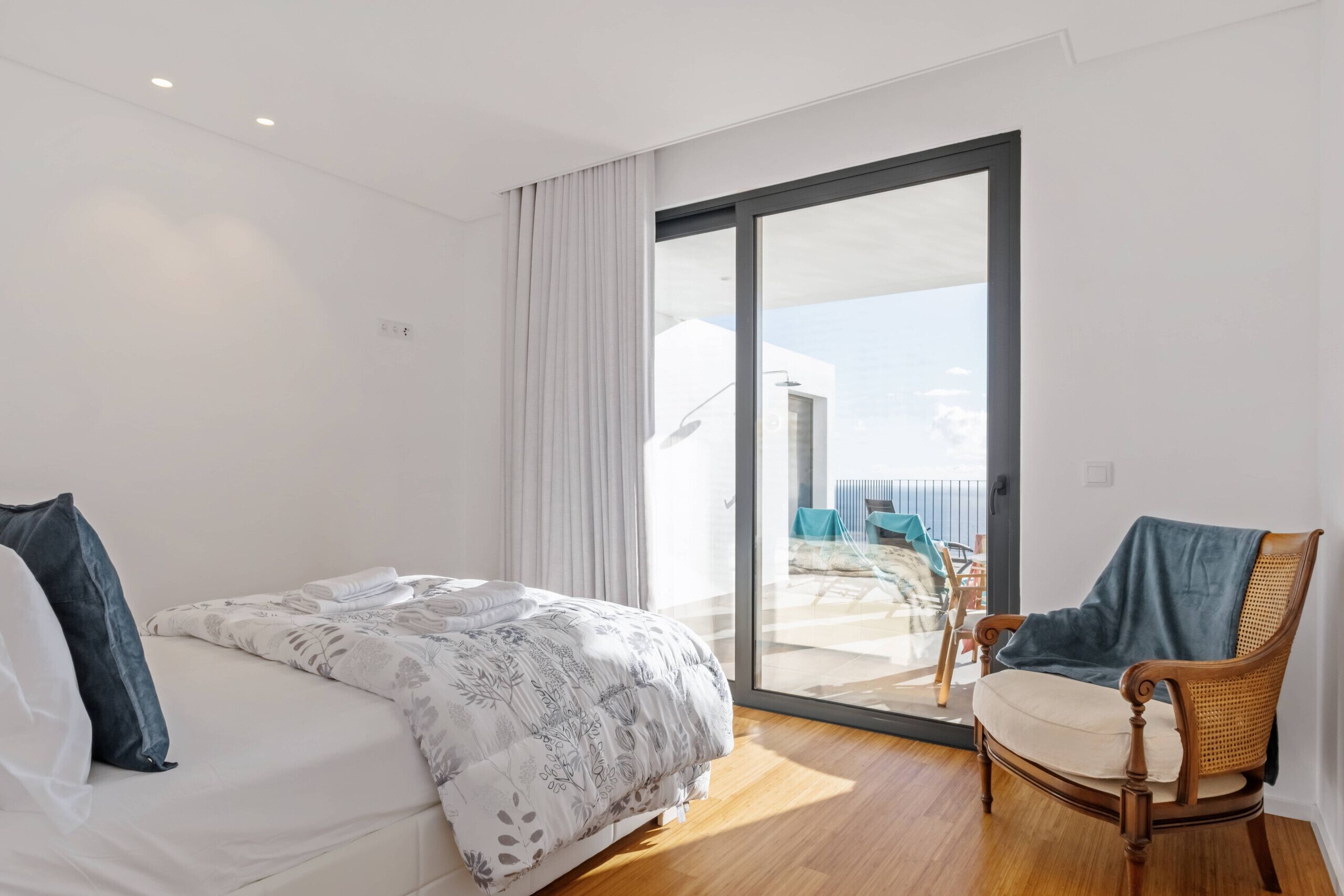 A modern hotel room with a large bed, a wooden chair, and a sliding glass door leading to a balcony with outdoor furniture.