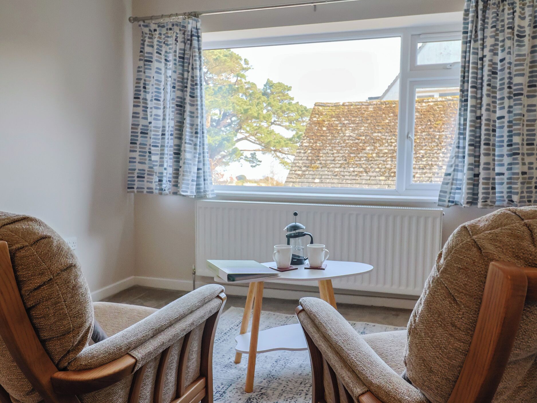 A room with two wooden chairs, a small table with a coffee pot, cups, and a book. A window with curtains is visible, and a radiator is below the window.