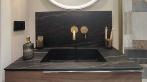 Un baño moderno con lavamanos de mármol negro, grifos dorados y un espejo grande con luz circular.