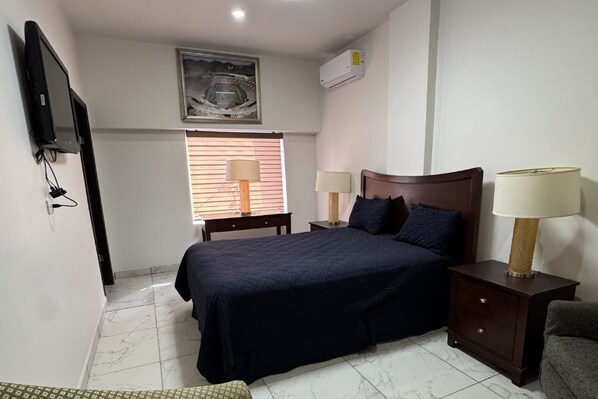 A bedroom with a bed, two bedside tables with lamps, a TV mounted on the wall, and a window with blinds. - Hotel La Frontera (Ciudad Juarez)