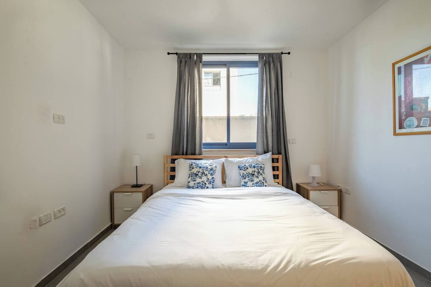 Een hotelkamer met een eenpersoonsbed, twee nachtkastjes, een raam met gordijnen en een aan de muur bevestigd schilderij.