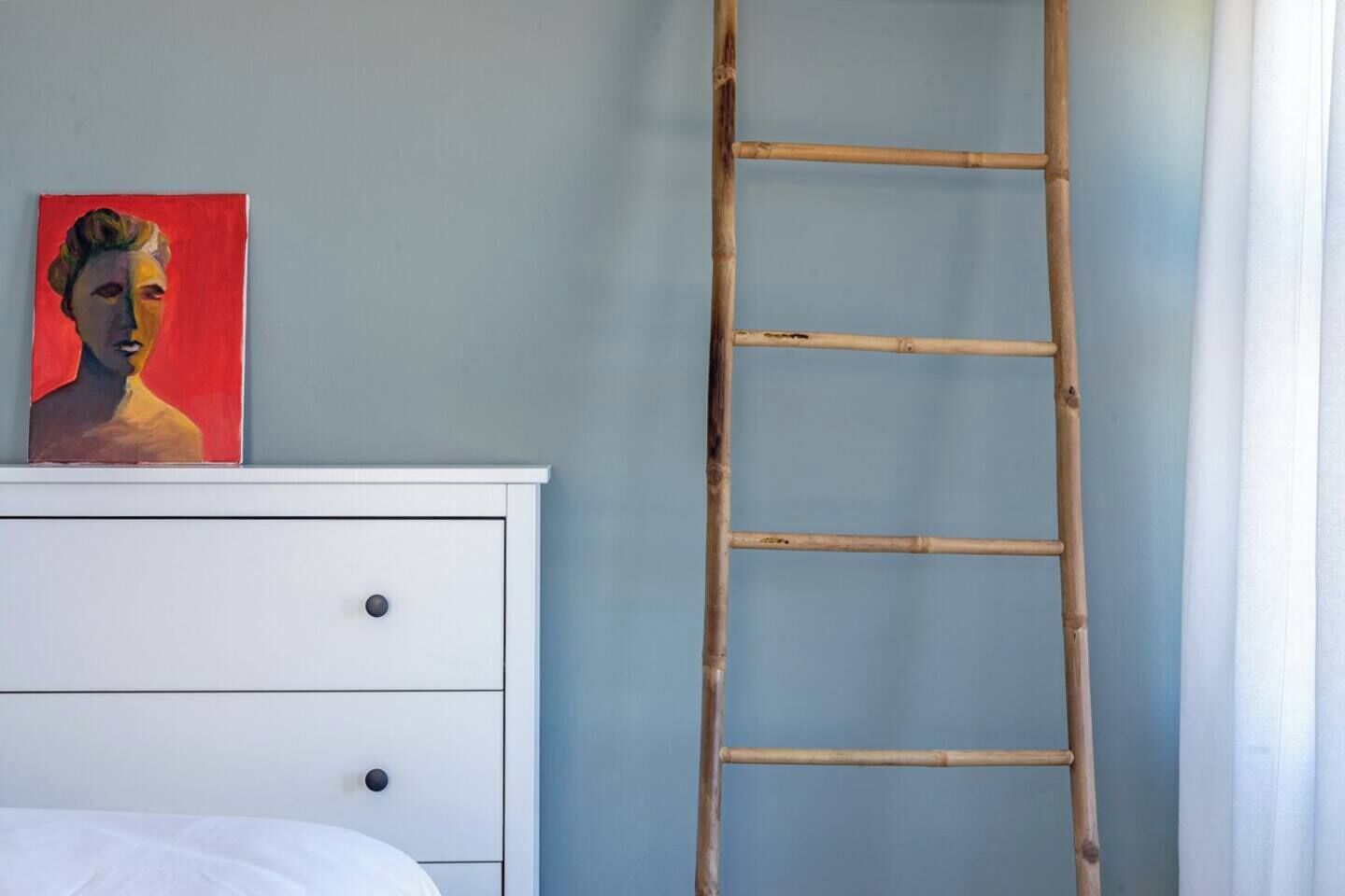 Una habitación con un aparador blanco, un cuadro encima y una escalera de bambú apoyada en una pared.