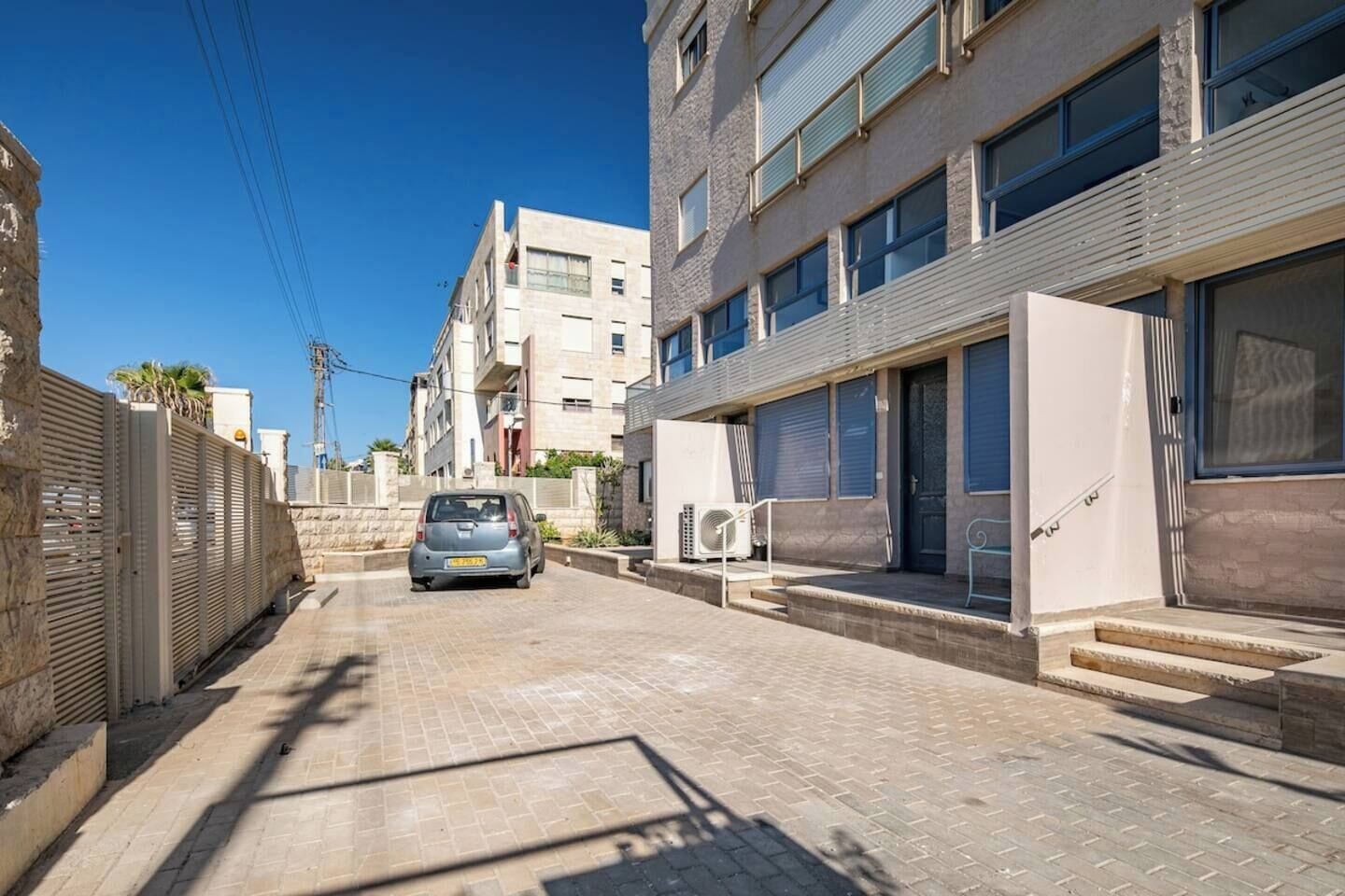 Una estrecha calle urbana con un coche estacionado, edificios con balcones y un cielo azul despejado.