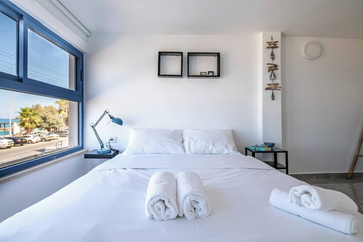 Une chambre d’hôtel comprenant un lit, deux tables de chevet, un bureau, une fenêtre donnant sur la rue et un luminaire fixé au mur.