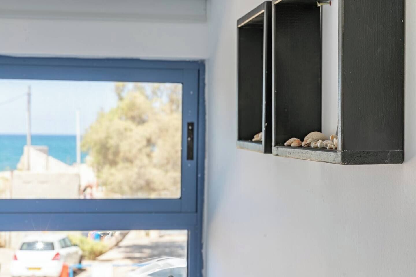 A room with a view of the sea through a window, a door, and a wall-mounted shelf with seashells.