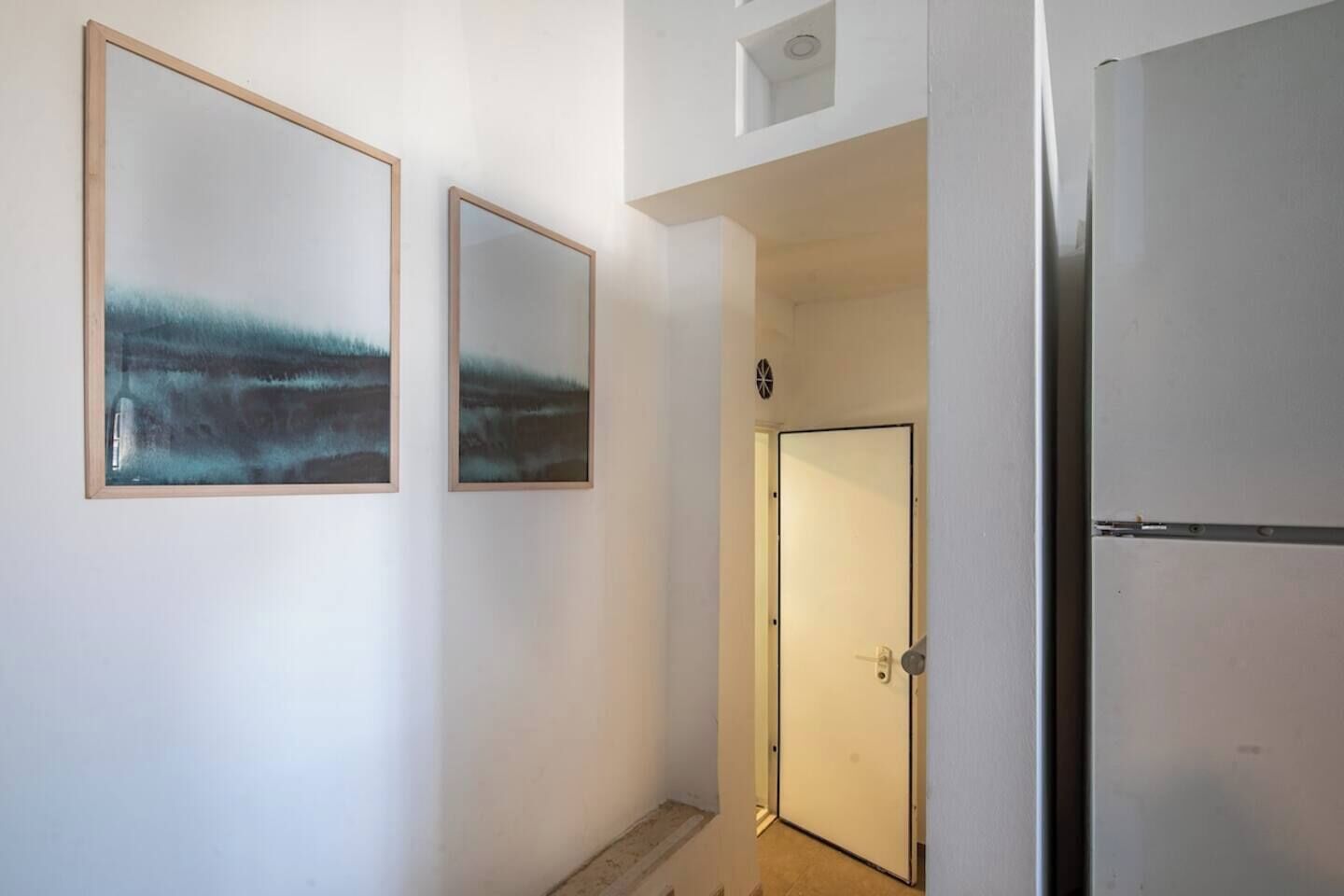 Um corredor com duas pinturas abstratas emolduradas na parede, uma porta com puxador e uma geladeira.