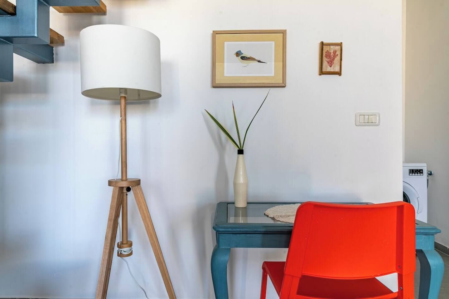Um quarto com uma luminária de chão, uma mesa com um vaso e uma moldura, uma cadeira vermelha e uma máquina de lavar.