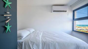 A bedroom with a bed, an air conditioning unit, a window with a view of the beach, and a wall decoration.