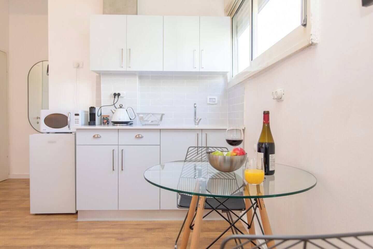 Una cocina moderna con armarios blancos, un microondas y una mesa de vidrio con un tazón de fruta y una botella de vino.