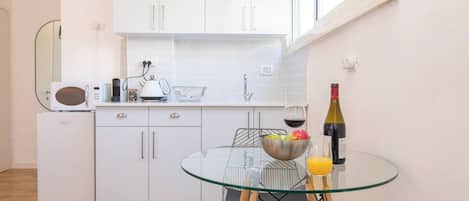 A modern kitchen with white cabinets, a microwave, and a glass table with a bowl of fruit and a bottle of wine.