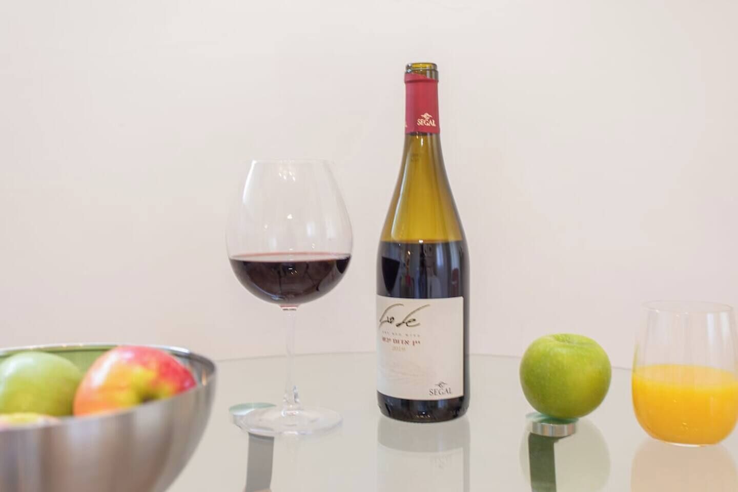 A wine bottle, a wine glass, a bowl of fruit, and a glass of juice on a reflective surface.