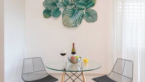 Un comedor con una mesa de vidrio, dos sillas, un frutero con fruta, una copa de vino y una botella de vino.