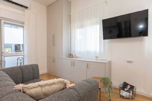 Een woonkamer met een grijze bank, een wit kastje, een flatscreen televisie en een raam met gordijnen.