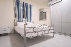 A single-bed room with a metal frame, white bedding, a bedside table with drawers, a window with blinds, and an air conditioning unit. - Port city haifa - 1 bdrm apt Near beach 2 (Haifa)