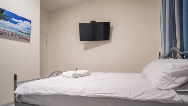 A single bed with white linens, a flat-screen TV mounted on the wall, and a painting of a beach scene. - Port city haifa - 1 bdrm apt Near beach 2 (Haifa)