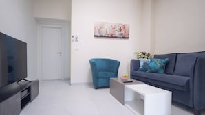 A modern living room with a flat-screen TV, a blue armchair, a grey sofa with decorative pillows, and a white coffee table. - Port city haifa - 1 bdrm apt Near beach 2 (Haifa)