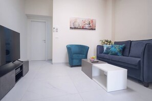 A modern living room with a flat-screen TV, a blue armchair, a grey sofa with decorative pillows, and a white coffee table. - Port city haifa - 1 bdrm apt Near beach 2 (Haifa)