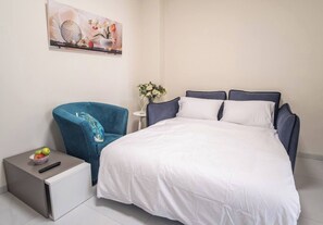 Quarto de hotel com cama, uma poltrona azul, uma mesinha com uma tigela de frutas e um quadro na parede.