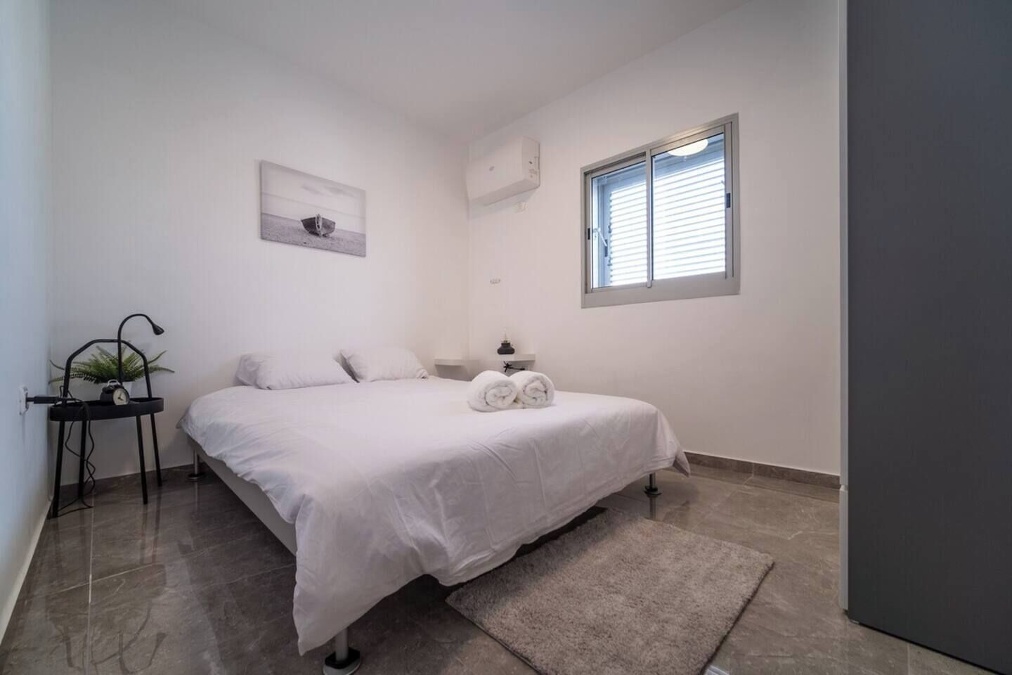 A bedroom with a bed, a bedside table, a lamp, a potted plant, a window, and a wall-mounted air conditioner.