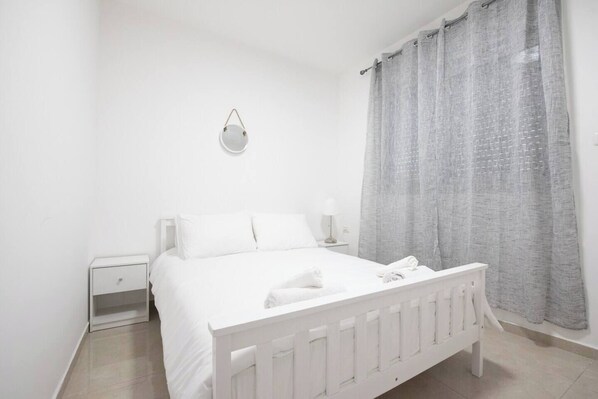 A white bedroom with a bed, nightstands, a wall-mounted light, and a window with grey curtains.