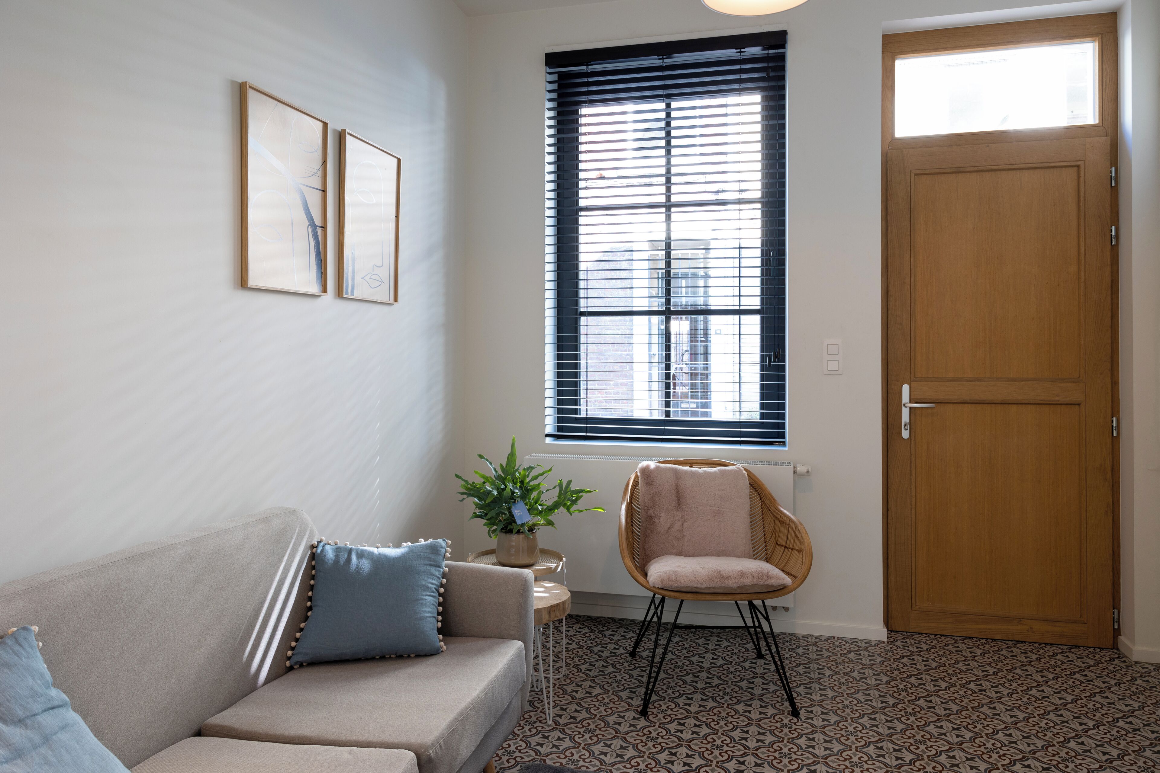 A living room with a beige sofa, a blue pillow, a potted plant, a wicker chair, and a wooden door.