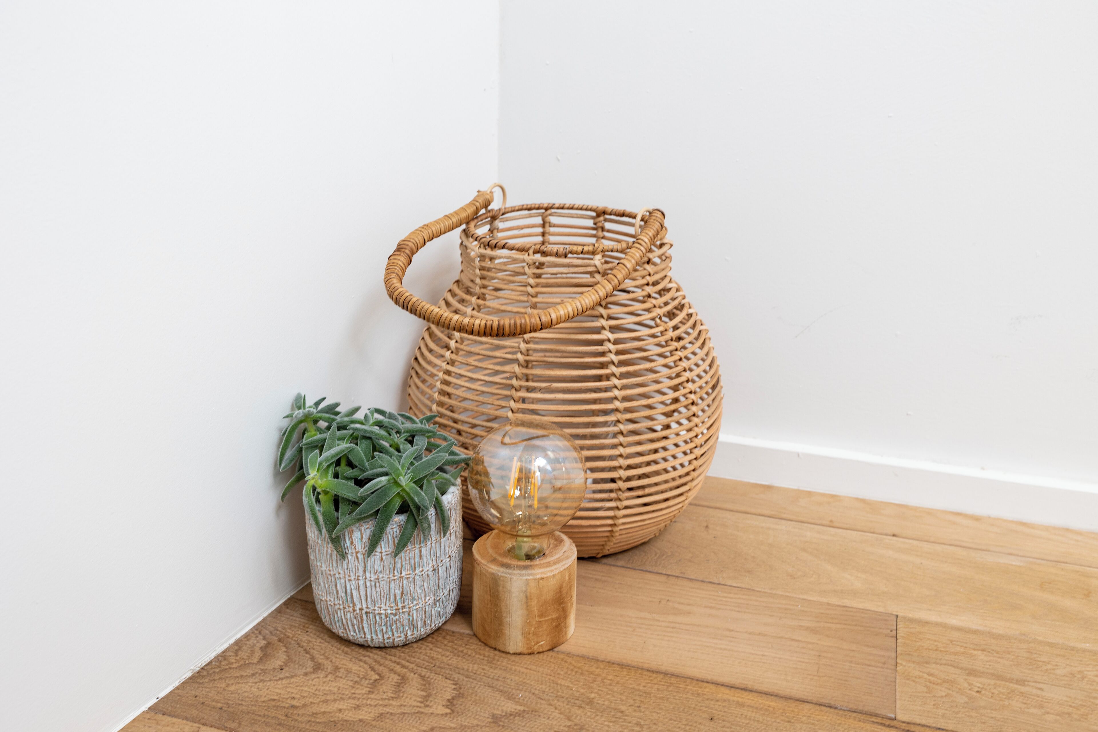 A wicker basket, a potted plant, and a glass object on a wooden floor against a white wall.