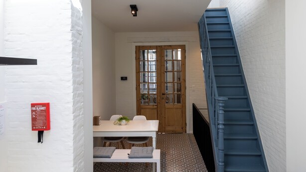 A narrow hallway with a white brick wall, a wooden door, a blue staircase, and a white table with chairs. - Huys17 (Hasselt)