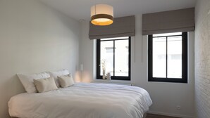 A bedroom with a large bed, two windows with blinds, a hanging light fixture, and a brick wall. - Huys17 (Hasselt)