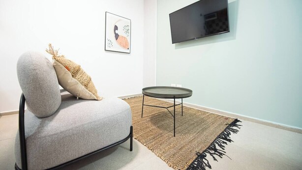 Een moderne woonkamer met een grijze bank, een klein rond tafeltje, een flatscreen televisie aan de muur en een kunstwerk in een lijst boven de bank.