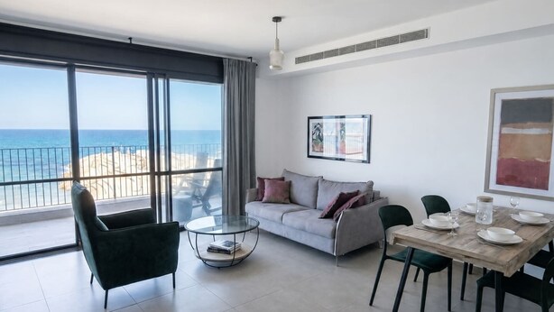 A modern living room with a balcony overlooking the sea, a dining area with a table set for two, and a sofa with cushions. - Port city- 1 bdrm apt w/ balcony beachfront+ mamad (Haifa)
