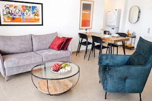 A living room with a grey sofa, a round glass coffee table with fruit, a blue armchair, and a dining area with a wooden table and chairs. - Port city- 1 bdrm apt w/ balcony beachfront+ mamak (Haifa)