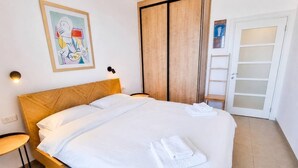 A neatly made bed with white linens and towels, a wooden headboard, a wall-mounted light, and a door with frosted glass panels. - Port city- 1 bdrm apt w/ balcony beachfront+ mamad (Haifa)