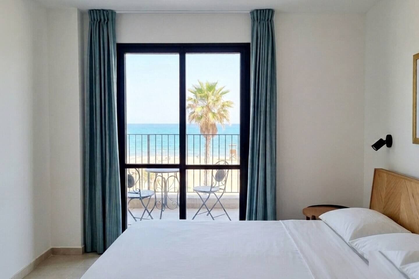 Habitación de hotel con cama, balcón con vista a la playa y palmeras, y una pequeña mesa con sillas.