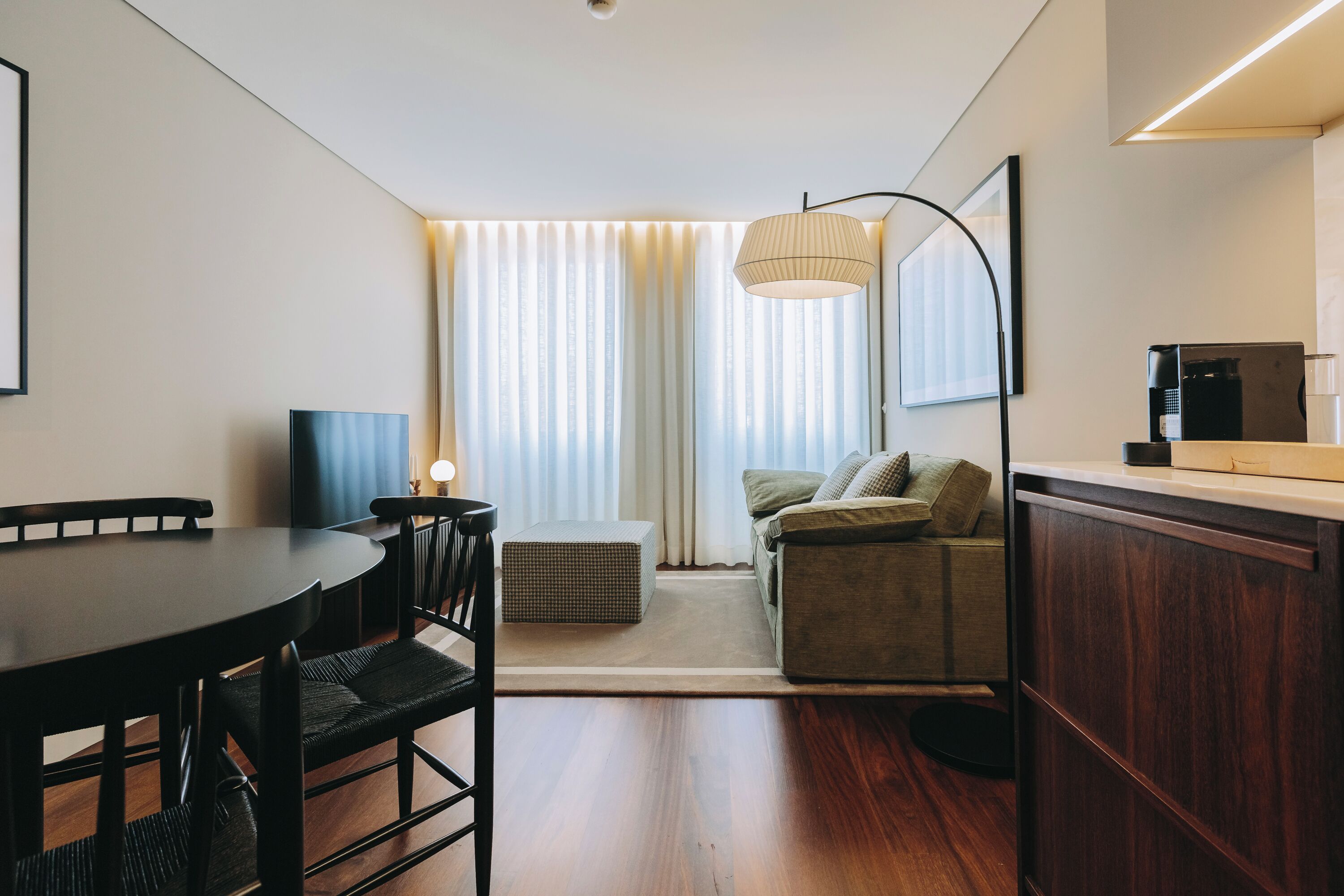 Ein modernes Hotelzimmer mit Essbereich, einer Couch, einer Kaffeemaschine und einem großen Fenster mit Vorhängen.