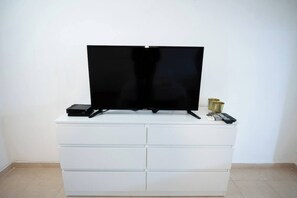 A flat-screen TV on a white cabinet with drawers, a remote control, and a small device on top. - Downtown Studio with gallery nxt german colony (Haifa)