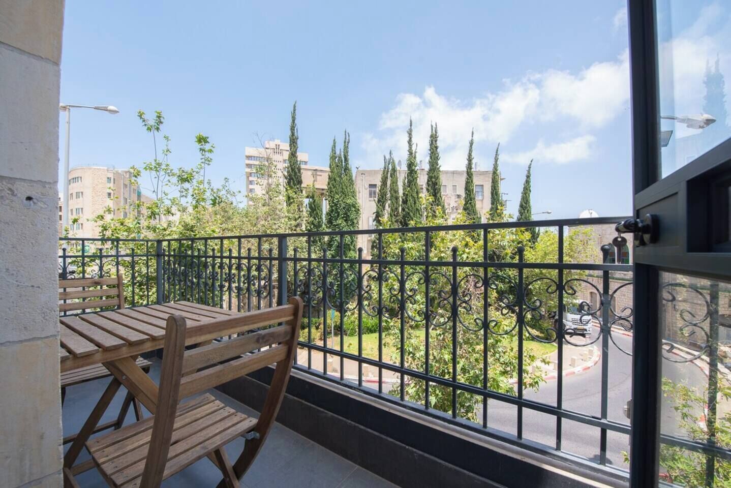 Un balcon avec une table et des chaises en bois, une rambarde en métal et une vue sur un paysage urbain avec des bâtiments et des arbres.