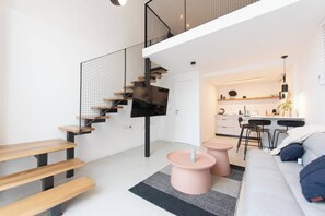 Eine moderne Wohnung mit minimalistischem Design, ausgestattet mit einer Treppe aus Holz und einem Metallgeländer, einem Wohnbereich mit einer Couch und zwei rosafarbenen Beistelltischen sowie einer Küchenzeile mit einer Bar und Hockern.