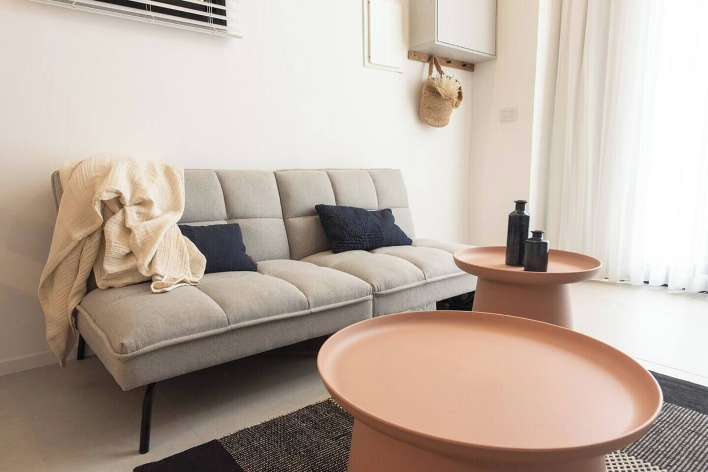 A modern living room with a sofa, two side tables, a hanging bag, and a window with curtains.