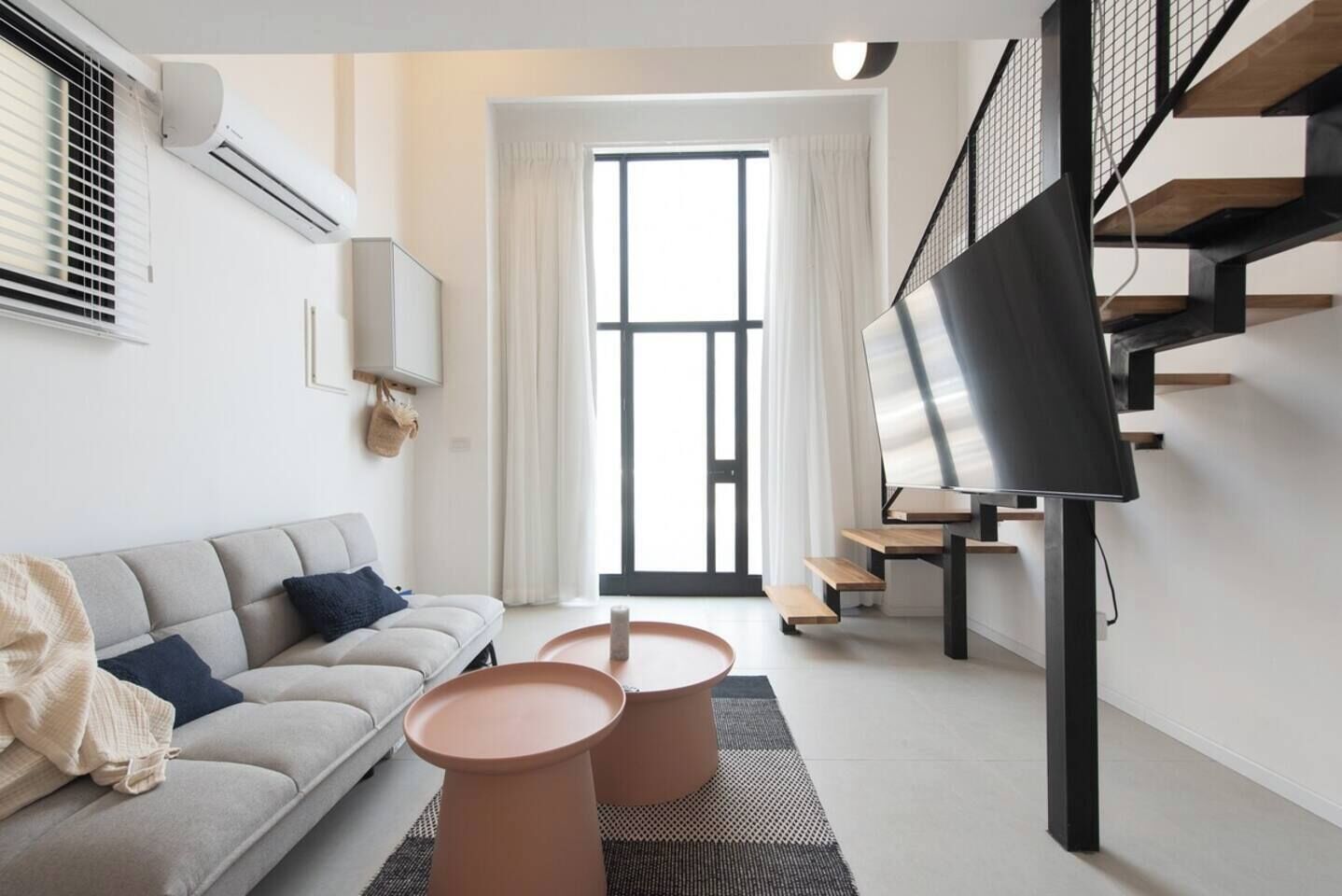 Una sala de estar moderna con un sofá, dos mesitas auxiliares de terracota, un televisor de pantalla plana sobre un mueble bajo, una escalera con barandilla de metal y un ventanal con cortinas.