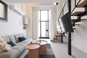 Ein modernes Wohnzimmer mit einer Couch, zwei Terrakotta-Beistelltischen, einem Flachbildfernseher auf einem Standfuß, einer Treppe mit Metallgeländer und einem großen Fenster mit Vorhängen.