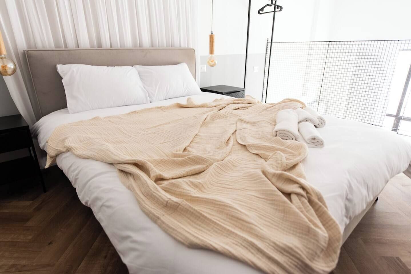 A neatly made bed with a beige blanket, white pillows, and rolled towels on the side.